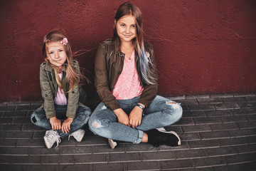 Two pretty smiling girls are sitting on the floor near red wall on the street.