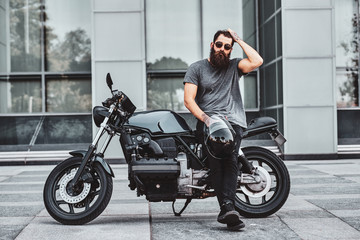 Brutal tattooed biker in sunglasses is resting next to his motorbike in the city centre.