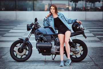 Beautiful young woman in minishorts and denim jacket is posing on the bike while holding helmet.