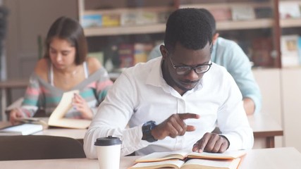 African american male college student in white shirt, wearing spectacles preparing for exams in library, reading a book at table.