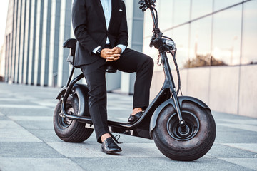 Fototapeta na wymiar Elegant man in black suit is sitting on his electro scooter while posing for photographer.
