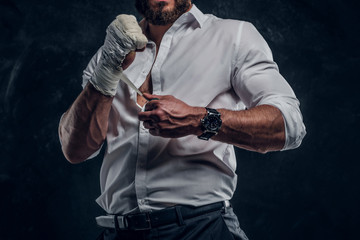 Bearded brutal man in white shirt is wearing protection on his fist before fight at dark studio.