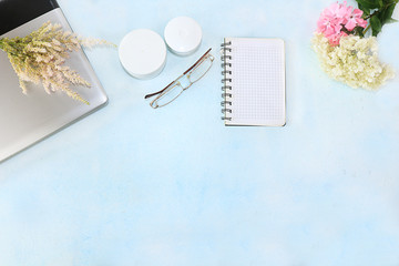Cream for skin care, a laptop on a sunny table, a notebook for work and glasses, multi-colored twigs of flowers, preparing for a summer vacation in the sun, copy space, top view. 