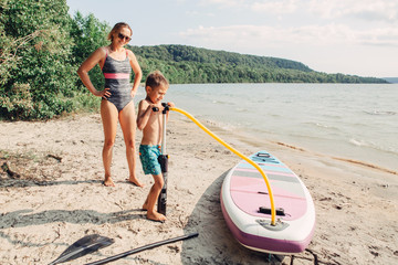 Caucasian woman mother teaching son to inflate sup surfboard with pump on beach. Happy family...