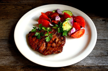 Pork steak, salad tomatoes, cucumbers greens seasonings on a white plate.