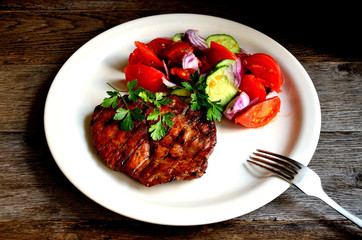 Pork steak, salad tomatoes, cucumbers greens seasonings on a white plate.