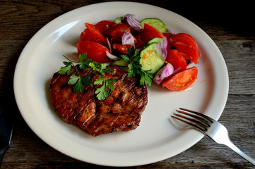 Pork steak, salad tomatoes, cucumbers greens seasonings on a white plate.