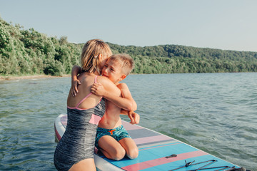 Fototapeta na wymiar Caucasian woman mother hugging comforting crying sad son boy child on paddle sup surfboard in water. Mom Modern outdoor family activity sport hobby.