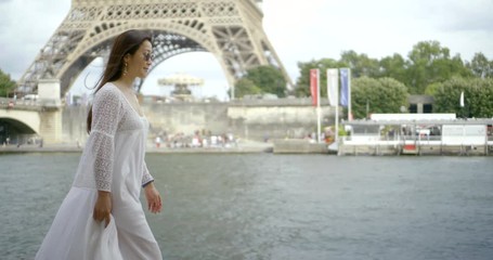 a dark-haired Asian middle-aged woman walks in Paris against a bridge, along the river Seine and in the background of the Eiffel tower. She's wearing a long white dress and dark glasses.