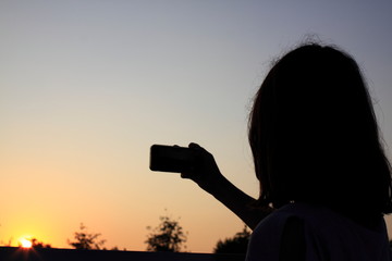 Silhouette of a young girl with a mobile phone. Teenager makes selfie on the phone against the sunset. 