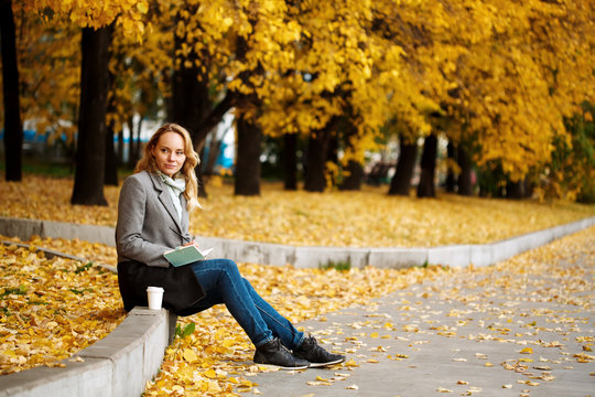 Woman sitting in autumn city park with golden trees,writing something in her notebook - Powered by Adobe