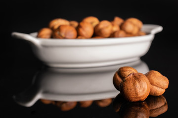Lot of whole ripe brown hazelnut in white oval ceramic bowl isolated on black glass
