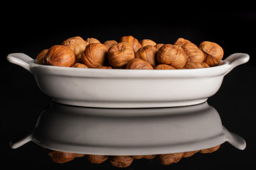 Lot of whole ripe brown hazelnut in white oval ceramic bowl isolated on black glass
