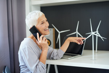 Gorgeous Caucasian senior woman talking on the phone and touching windmill model while sitting in office. Start up business concept.