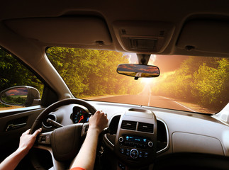 Car dashboard with driver's hands on the steering wheel and rear view mirrors on a road with trees against sky with sunset