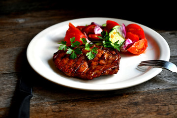 Pork steak, salad tomatoes, cucumbers greens seasonings on a white plate.