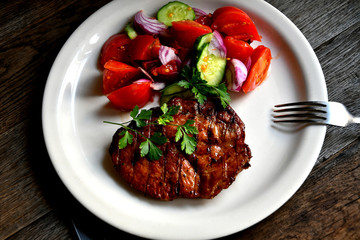 Pork steak, salad tomatoes, cucumbers greens seasonings on a white plate.