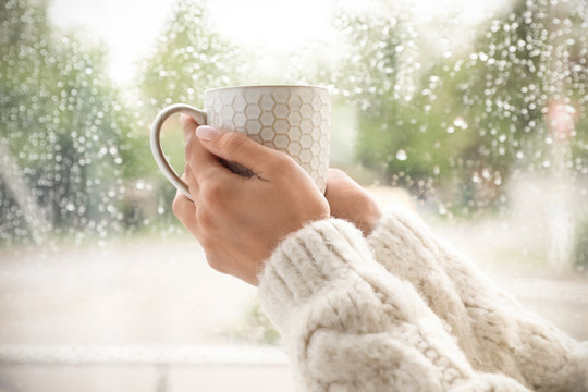 Young woman with cup of tea near window indoors on rainy day, closeup - Powered by Adobe
