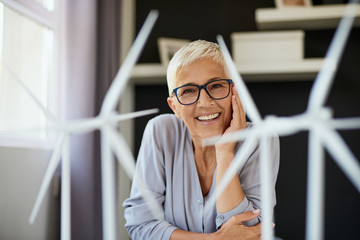 Happy Caucasian senior woman leaning on desk and looking at camera. On desk are windmill models....