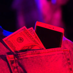 Hundred dollar bill and smartphone in pocket of blue jeans, toned. Dollars in neon light. Banknotes of 100 and 50 american dollars and mobile phone in denim jeans pocket