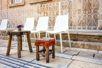 A beautiful flower stands on a table made of wood cut. Next to the wooden table is a small wooden chair and several white plastic chairs. A cozy place to relax on the street of the beautiful city.
