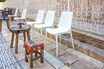 A beautiful flower stands on a table made of wood cut. Next to the wooden table is a small wooden chair and several white plastic chairs. A cozy place to relax on the street of the beautiful city.