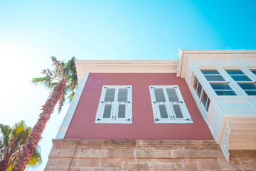 A palm tree next to the house is pink. The house has two large white windows and a balcony. House and palm tree against a clear blue sky.