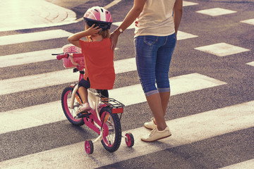   Mother goes pedestrian crossing with daughter on bicycle. A woman with child crossing the road in...