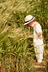 Naklejka na ściany i meble A cute beautiful caucasian boy dressed in a linen cloth and white hat standing in a field of green wheet