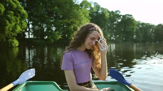 Attractive curly girl sitting in a boat and smiling. Happy young woman in a purple t-shirt holds glasses in her hands on a background of green landscape. Summer outdoor activities.