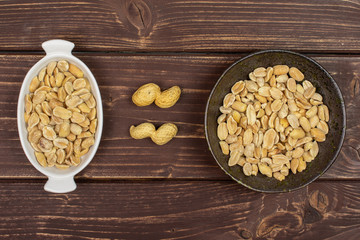 Lot of whole lot of halves of natural yellow peanut in white oval ceramic bowl in dark ceramic bowl flatlay on brown wood