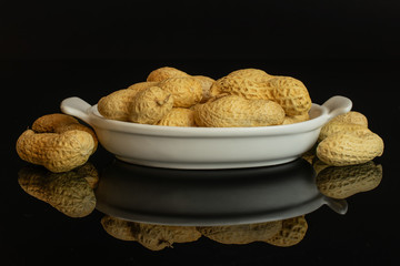 Lot of whole natural yellow peanut in white oval ceramic bowl isolated on black glass