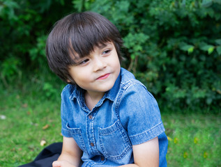 Portrait of a young boy sitting  on lawn in the park, Active child looking out with smiling face sitting on green grass field, Kid having fun playing outdoors in sunny day summer,
