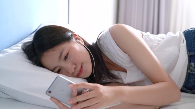 Beautiful Asian woman listening music with smartphone smiling happy in the bed her lying under blanket in bedroom comfortable resting hotel people lifestyle Slow motion 4k resolution.