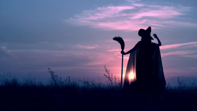 back view view sorceress in fluttering cloak and hat holding broom posing on field on the background a blue sky with clouds all saints night