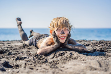 Black Friday concept. Smiling boy with dirty Black face sitting and playing on black sand sea beach...