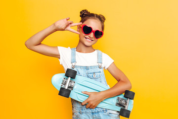 Happy teenager with skateboard, stylish girl posing in sunglasses on yellow background