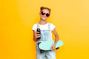 Happy teenager, girl in sunglasses with a skateboard and a refreshing drink in hand, relaxing on a yellow background