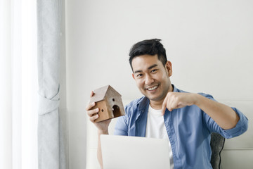 Asian man holding house in his hand and using laptop, real estate concept.