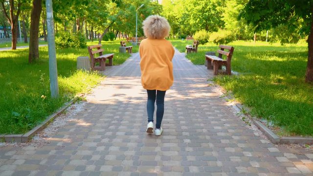 back view woman with curly blond hair wearing jeans and yellow sweater walking in summer park nature outdoors background