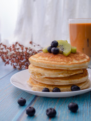 Stack of pancakes with topping, kiwi and blueberry.placed in a white plate on a blue wooden table.Eat with milk tea in the glass.