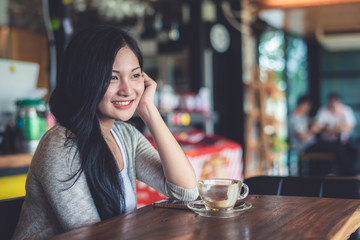Beautiful attractive young Asian woman holding a cup of coffee in hand and sitting on sofa at cafe in the morning, vintage color tone. 