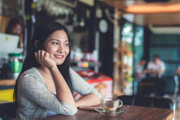 Beautiful attractive young Asian woman holding a cup of coffee in hand and sitting on sofa at cafe in the morning, vintage color tone. 
