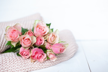 Obraz na płótnie Canvas Pink roses laying on a thick knit woolen cosy blanket laying on a white wooden background 