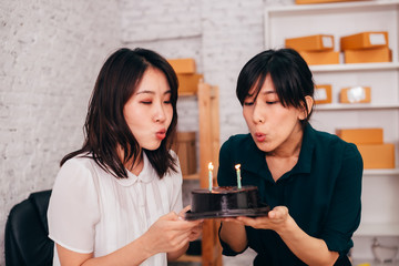 Cheerful Asian colleagues smiling and blowing candles on cake while celebrating birthday in office