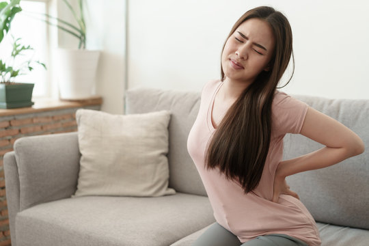 Asian girl having Waist pain sitting on sofa at home. young girl  backache from the muscles, Health and illness concepts