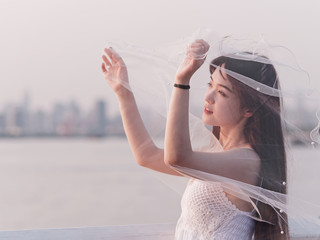 Portrait of beautiful Chinese girl in white bride dress looking away with veil in hand in windy dusk with blur river water background, Beauty, lifestyle and summer Concept.