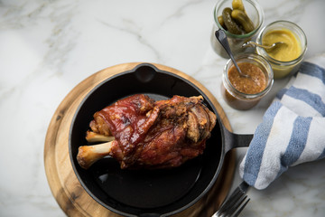 Gegrillte Schweinefleisch Haxe in gusseiserne Pfanne auf Holzbrett mit Cornichons, süßen und mittelscharfen Senf auf Marmor Hintergrund