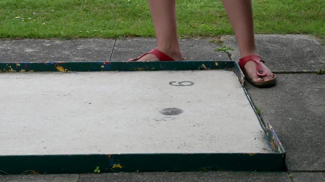 Eine Frau in roten Sandalen auf Minigolfplatz schl&auml;gt den Minigolfball. Der Ball rollt &uuml;ber die Bahn. Die Frau geht weg