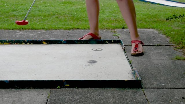 Eine Frau in roten Sandalen auf Minigolfplatz schl&auml;gt den Minigolfball. Der Ball rollt &uuml;ber die Bahn. Die Frau geht weg